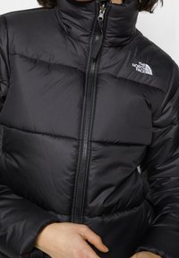 Chaqueta acolchada negra con cuello alto, cierre de cremallera y paneles cosidos. Presenta un pequeño logotipo en el lado izquierdo y textura suave.