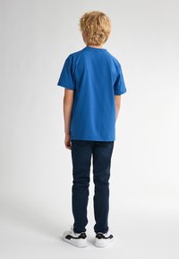 Camiseta de algodón azul con mangas cortas, cuello redondo y corte relajado, combinada con pantalones de mezclilla oscuros y zapatillas blancas con detalles en negro.