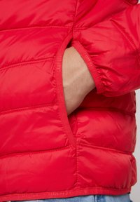 Rote Steppjacke mit quadratischem Design, ausgestattet mit einer Seitentasche, elastischen Bündchen und glatter Textur. Im Inneren ist ein Markenetikett zu sehen.