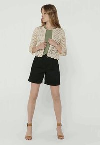 Woman wearing a beige crochet cardigan over an olive green top, black shorts, and brown heeled sandals, standing against a plain background.