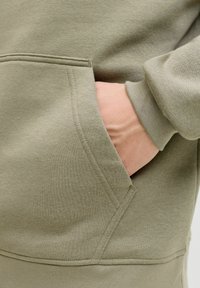 Dettaglio di una felpa grigio-verde con una tasca frontale in cui si intravede una mano posata. Texture morbida e liscia con cuciture visibili attorno alla tasca.