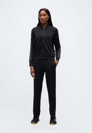 Femme debout, portant une veste de sport noire zippée et un pantalon assorti avec des bandes sur les manches et les jambes, baskets noires.