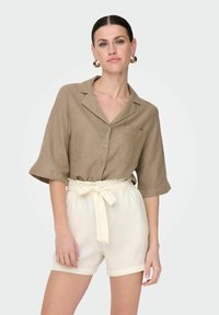 Camicia di lino marrone a maniche corte, con colletto e tasca frontale, abbinata a pantaloni corti color crema con vita regolabile e orlo arrotolato.