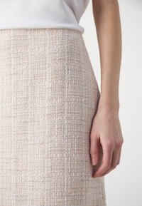 Gros plan sur une personne portant une jupe taille haute beige texturée et un haut blanc, avec le bras droit reposant le long du corps.