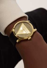 Montre Guess couleur or avec un cadran triangulaire et un motif en maille, dotée d'un bracelet en silicone noir et d'une couronne proéminente.