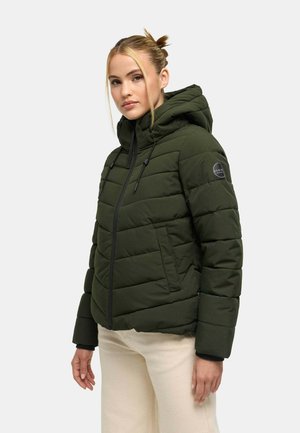 Frau mit blondem Haar, die eine dunkelgrüne Steppjacke mit Kapuze und beige Hose trägt, steht vor einem schlichten hellen Hintergrund.