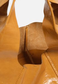 Primer plano del interior de una bolsa de cuero color tostado que muestra el forro de ante y los bordes cosidos con el nombre de la marca grabado en la superficie interna.