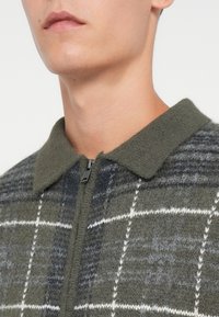 Casaco em mistura de lã com padrão xadrez em cinza e verde, com fecho de zipper e um colarinho macio e texturizado.