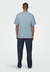 Helles, blaues Baumwoll-T-Shirt mit kurzen Ärmeln, lockere Passform; kombiniert mit dunkelblauen Jeans und weißen Turnschuhen. Schlichte Gestaltung, ohne Muster.