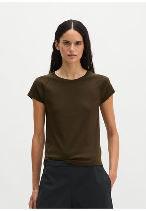 Donna con lunghi capelli scuri che indossa una maglietta attillata verde oliva scuro a maniche corte e pantaloni scuri, in piedi davanti a uno sfondo chiaro e semplice.