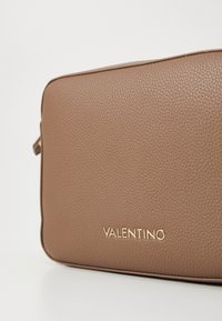 Bruine gestructureerde leren tas met een rechthoekige vorm, met een gouden "VALENTINO" logo aan de voorkant. Zachte ritsduur zichtbaar aan de zijkant.