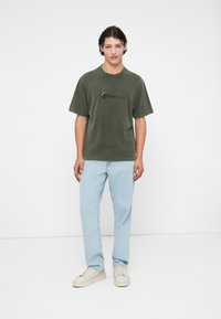 Olijfgroen oversized T-shirt met korte mouwen, met een grafische print op de borst. Gepaard met lichtblauwe denim en witte sneakers.