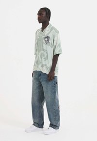 Short-sleeve light green button-up shirt with abstract patterns, paired with loose-fit faded blue jeans and white sneakers.