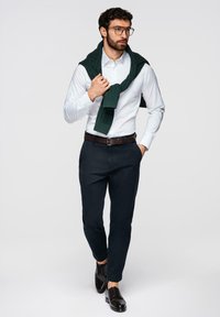 Homme à barbe et lunettes portant une chemise blanche, un pantalon bleu marine, des chaussures noires et un pull vert foncé drapé sur les épaules, marchant en avant.