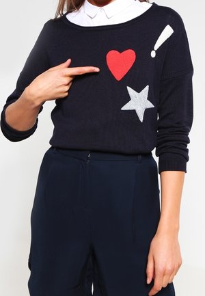 Persoon draagt een zwarte trui met rode hart-, witte ster- en witte uitroeptekenpatches, wijzend met de linkerhand naar het hart.