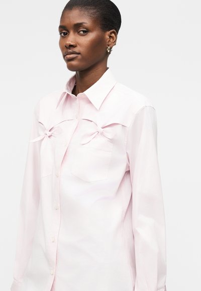 Femme aux cheveux courts portant une chemise rose pâle boutonnée avec des nœuds attachés sur les poches poitrine, debout devant un fond blanc uni.