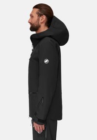 Schwarze wasserdichte Jacke mit Kapuze, ausgestattet mit Reißverschlusstaschen, elastischen Bündchen und einem Logo-Patch am Ärmel. Glatte Textur und modernes Design.