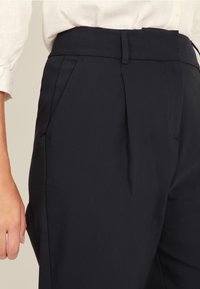 Närbild av en person klädd i högmidjade svarta skräddarsydda byxor och en inskjuten vit långärmad blus, med handen avslappnat vid sidan.