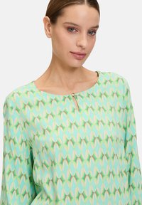 Blusa leggera con uno sfondo verde menta che presenta un motivo geometrico ripetitivo in tonalità di verde, beige e marrone. Dettaglio con scollatura a chiave.