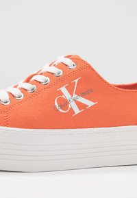 Orange canvas sneaker med vita snören och en texturerad ovandel. Har en vit logotyp och en tjock vit sula med horisontella linjer.