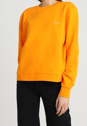 Kobieta ubrana w jasnopomarańczową bluzę Calvin Klein i czarne spodnie, z pierścionkiem na lewej dłoni, stoi na tle jednolitego tła.