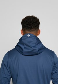 Veste à capuche bleu marine en tissu lisse, avec un col montant, des coutures apparentes et un logo au niveau de la nuque au dos.