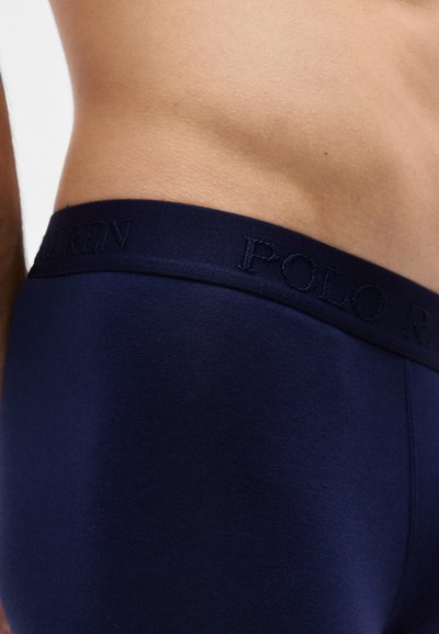 Ανδρικός κορμός από το πλάι, φοράει εσώρουχο Polo Ralph Lauren σε ναυτικό μπλε με επώνυμη ζώνη, σε απλό φόντο.