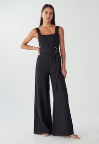 Žena s dlouhými tmavými vlasy, oblečená v černém bezrukávovém overal s opaskem a béžovými sandály, stojící s jednou rukou na boku.