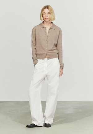 Donna con capelli biondi corti che indossa un maglione beige con bottoni, pantaloni bianchi a gamba larga e scarpe nere, in piedi con una mano in tasca.