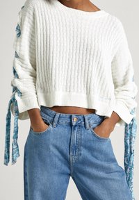 Suéter blanco de canalé con cintas azules estampadas en las mangas, combinado con jeans de mezclilla azul de cintura alta, con las manos en los bolsillos.