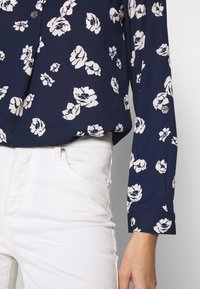 Námořnická květinová blůza s bílými květinovými vzory, dlouhými rukávy a detailem knoflíků, kombinovaná s bílými kalhotami, které ukazují hladkou tkaninu a uvolněný střih.