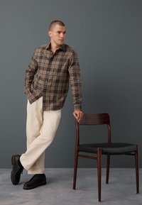 Camicia a quadri marrone e beige con bottoni, pantaloni color crema, scarpe nere. Appoggiato a una sedia di legno con seduta nera, su uno sfondo grigio.
