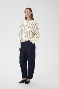 Femeia poartă un cardigan alb cu dungi verticale negre și nasturi maro, pantaloni largi bleu închis și balerini negri.