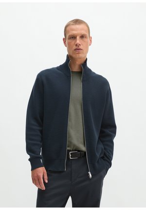 Mann trägt einen dunkelblauen Zip-Up-Pullover über einem olivgrünen Hemd mit dunkler Hose und schwarzem Gürtel vor schlichtem hellem Hintergrund.