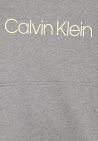 Grå bomullshoodie med texten "Calvin Klein" i ljusgul. Inkluderar en framficka med en mjuk textur och en enkel design.