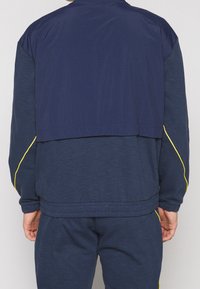 Námořnicky modrá sportovní bunda z texturovaného materiálu, s krátkým střihem, žlutými akcenty na rukávech a žebrovaným lemem.