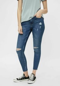 Jeans de mezclilla azul ajustados con detalles rasgados, con una cintura de altura media, bolsillos frontales y un silueta entallada, combinados con zapatillas negras.