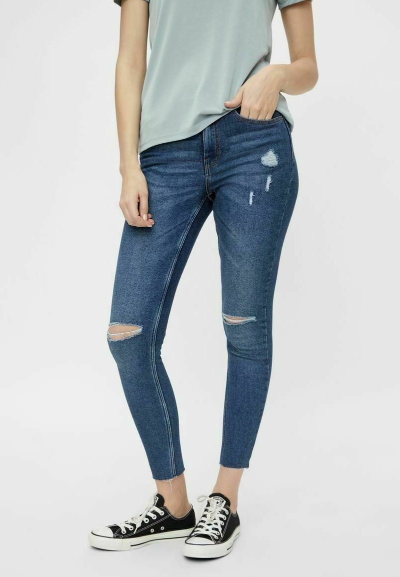 Jeans de mezclilla azul ajustados con detalles rasgados, con una cintura de altura media, bolsillos frontales y un silueta entallada, combinados con zapatillas negras.