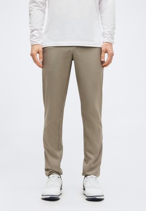 Άτομο που φοράει μπεζ παντελόνι ίσιας γραμμής, λευκό μακρυμάνικο πουκάμισο και λευκά παπούτσια με κορδόνια, πάνω σε απλό φόντο.