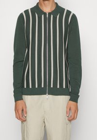 Grön zip-up kofta med vertikala beige ränder, ribbade ärmslut och nederkant. Har en klassisk krage och stickad textur.