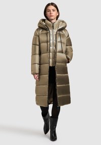 Palton pufos lung de culoare verde măslin, cu glugă, având un model matlasat, fermoar frontal și șnururi de strângere. Purtați deasupra unor pantaloni negri și cizme.