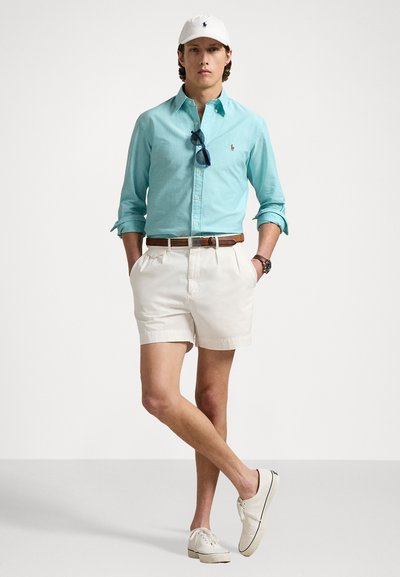 Gaiši zila krekla ar pogām, baltas šortus, brūnu jostu un baltas tenisa kurpes. Aksesuāri ietver saulesbrilles un baltu cepuri.