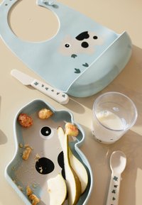 Silikoonist põll lehma kujutisega, sinine karu kujuline taldrik toiduga, lusikas ja tass piimaga, asetatud heledale pinnale.