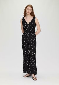 Mono negro con patrones abstractos en blanco, escote en V, diseño sin mangas, cintura anudada, piernas anchas, acompañado de sandalias negras con tiras.