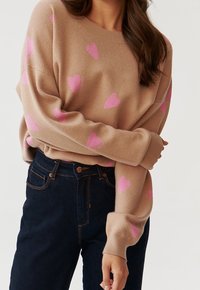 Pull en maille beige avec motifs de cœurs roses, encolure ronde et manches longues, associé à un jean denim foncé. Texture douce, coupe décontractée.