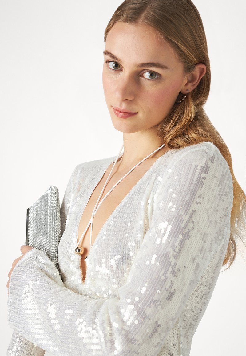 Biała sukienka z cekinami, z głębokim dekoltem w kształcie litery V i długimi rękawami; modelka trzyma srebrną kopertówkę; posiada długi biały sznurek z metalowym akcentem.