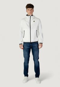 Giacca leggera bianca con zip frontale, dettagli neri e tasche laterali, abbinata a jeans blu scuro e sneakers bianche.