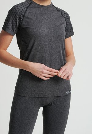 Femme portant un haut de sport gris foncé ajusté et des leggings, debout les mains jointes à la taille devant un fond uni.