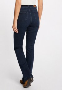Jean flare en denim foncé à taille haute, avec deux poches arrière et une texture subtile. Porté avec des bottes à talons marron.