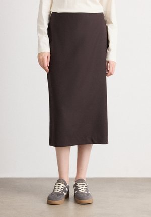 Personne portant une jupe midi marron foncé, un haut à manches longues crème et des baskets en daim gris avec des lacets blancs, debout sur un sol lisse.